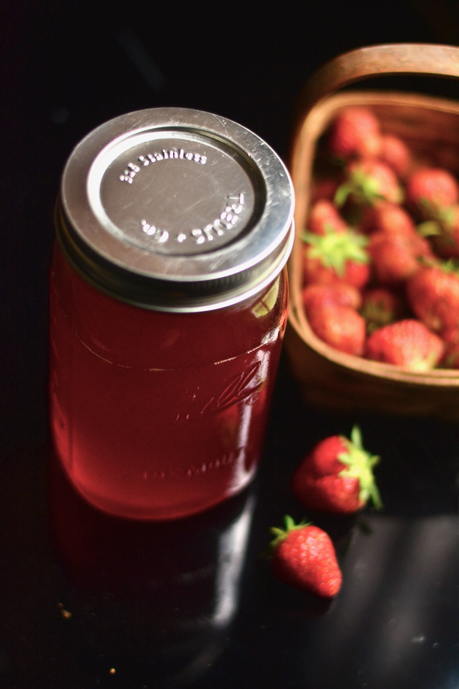 red syrup in jar on black surface with basket of just picked strawberries to the side