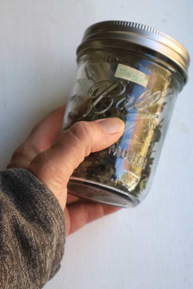 a hand holding dried basil leaves in a mason jar labeled with masking tape